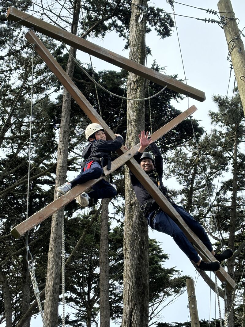 The image shows two people on a wooden structure in a forest. They appear to be partaking in an outdoor adventure activity, possibly a ropes course or climbing activity. Both individuals are wearing safety helmets and harnesses, indicating a focus on safety. The person on the right is waving at the camera.